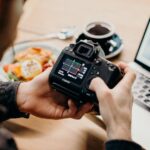 A man reviews photo settings on a camera while working on a laptop in a cozy workspace.