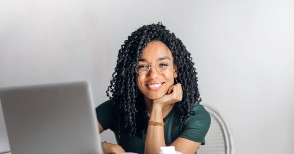 Joyful businesswoman with curly hair smiling at camera while using laptop indoors.