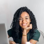 Joyful businesswoman with curly hair smiling at camera while using laptop indoors.