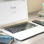 A neat workspace featuring a laptop displaying Google search, a smartphone, and a notebook on a wooden desk.